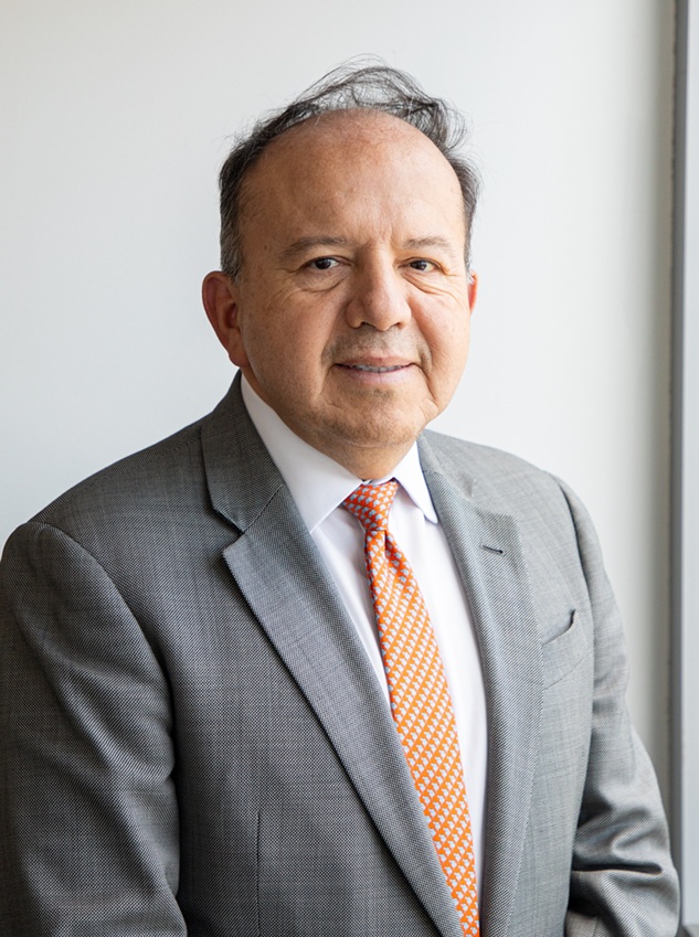 Professional headshot of a person wearing a gray business suit, white dress shirt, and orange patterned tie. The portrait is framed from the chest up against a light gray background, with the subject angled slightly toward the camera. Soft natural lighting creates a clean, polished appearance suitable for a professional profile or organizational directory.