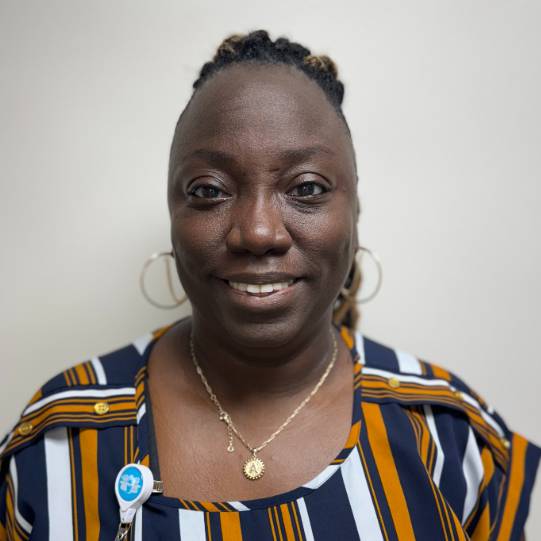 Professional portrait of a person standing against a plain light-colored wall. The person is wearing a navy, white, and gold vertically striped blouse, large hoop earrings, and a pendant necklace. An employee identification badge is clipped to the blouse near the shoulder. The image is evenly lit and framed from the chest up, with the person facing the camera.
