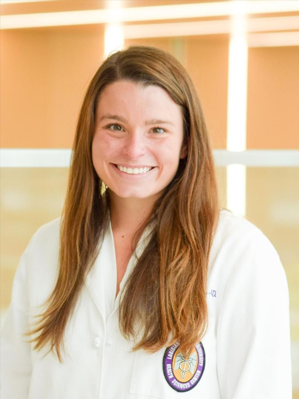 Professional medical school portrait of a person wearing a white medical coat against a bright indoor background with large windows.