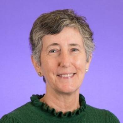 Professional head-and-shoulders portrait of a person with short gray hair wearing a green knit sweater with a ruffled mock neckline and small stud earrings. The person is facing the camera against a smooth purple studio-style background with soft, even lighting.