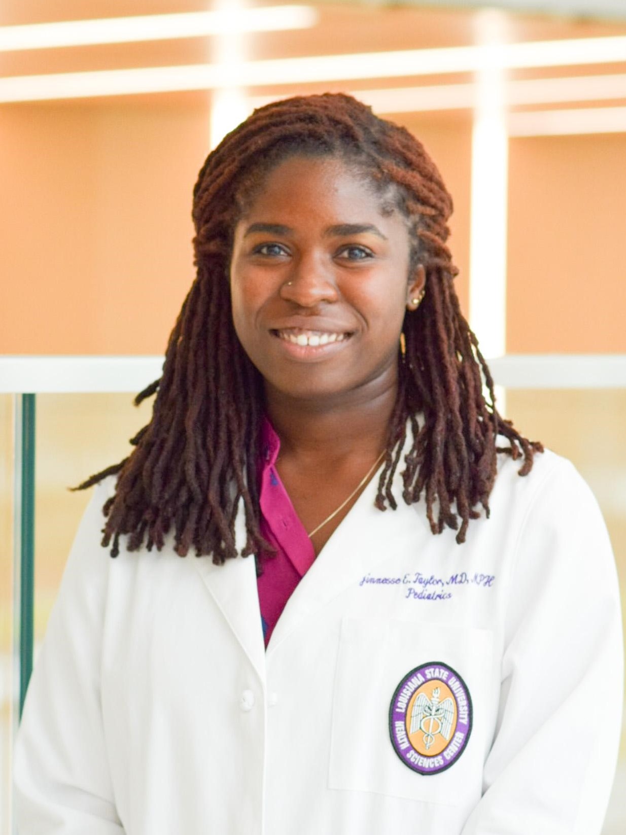 Professional medical portrait of a person wearing a white medical coat over a bright pink top, photographed indoors against a softly lit background with large window panels.