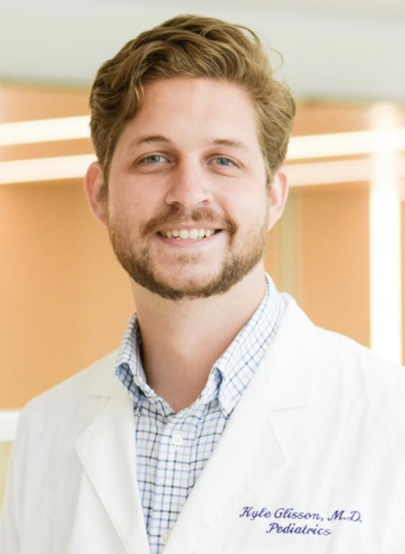 Head-and-shoulders professional portrait of a person wearing a white lab coat over a light blue collared shirt. The person is facing the camera with a slight smile. The background is softly blurred with warm beige and orange tones, creating a clean healthcare or clinical office setting. A small logo is visible on the lab coat but is not legible.