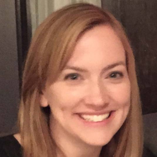 Close-up indoor portrait of a person with shoulder-length light brown hair wearing a black top. The person is facing the camera with a visible smile. The background is softly lit and out of focus, showing dark and neutral-colored interior elements.