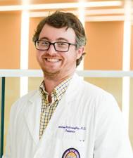 Professional headshot of a person wearing glasses, a white lab coat, and a checkered button-down shirt. The lab coat features embroidered text and a Louisiana State University Health Sciences Center patch near the lower pocket. The person is standing indoors in front of large windows with warm natural light and a softly blurred background.