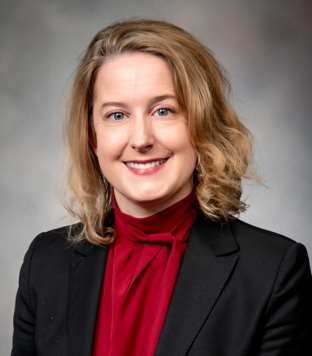 A professional studio headshot of a person with shoulder-length light brown hair, wearing a dark blazer over a red blouse with a tied neckline. The person is facing forward and smiling, set against a neutral gray background.