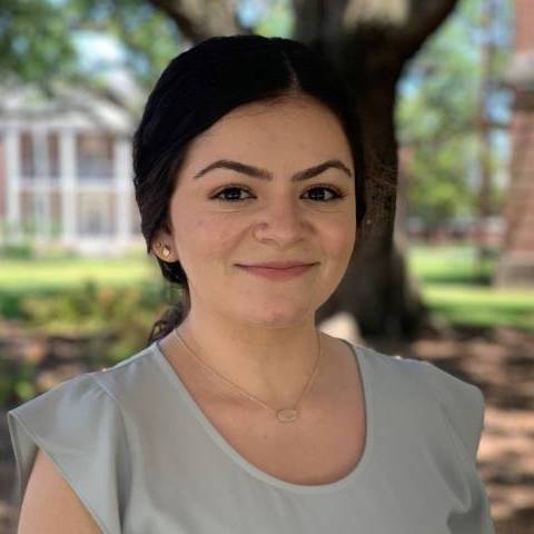 Outdoor portrait of a person standing beneath a large tree on a landscaped campus. The person has dark hair pulled back and is wearing a light gray short-sleeved blouse, small stud earrings, a delicate necklace, and light-colored pants. The background is softly blurred, showing trees, grass, and brick buildings in daylight.
