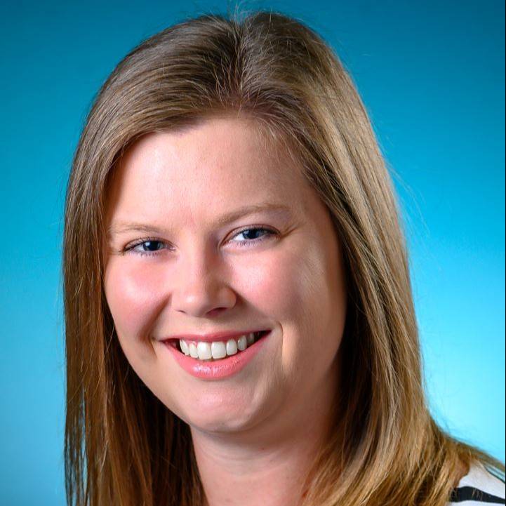 Professional studio headshot of a person with long, straight light brown hair wearing a white top with black horizontal stripes. The person is facing the camera against a bright blue gradient background. The portrait is closely cropped and evenly lit, creating a polished and professional appearance.