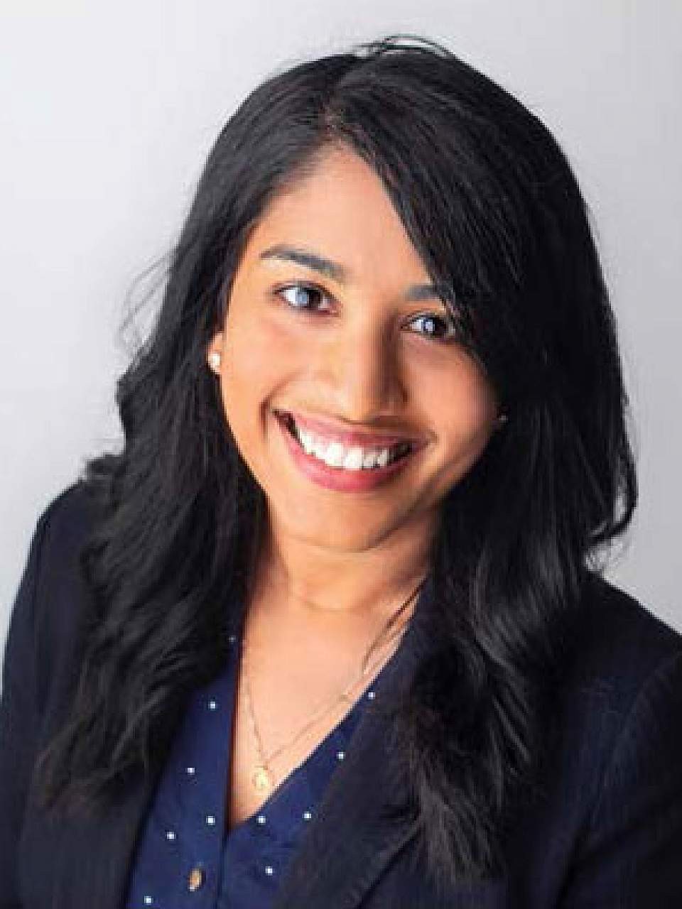 Professional headshot of a person with long, dark hair, wearing a dark blazer over a navy blouse with a subtle dotted pattern. The person is facing the camera with a visible smile. The portrait is cropped from the chest up and set against a plain light gray background.