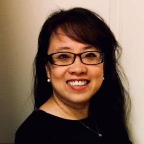 Head-and-shoulders portrait of a person with long dark hair and rectangular glasses, wearing a black top, pearl stud earrings, and a delicate necklace. The person is facing the camera with a visible smile, standing indoors against a light-colored wall and door background.