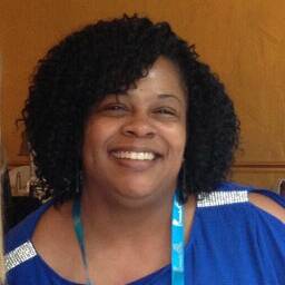 Head-and-shoulders portrait of a person with short, curly hair wearing a bright blue top with shoulder cutout details and a blue lanyard. The person is facing the camera indoors, with a wood-paneled wall and softly lit background visible behind them.