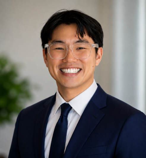 Professional headshot of a person wearing clear-framed glasses, a navy blue suit jacket, a white dress shirt, and a dark blue tie. The person is facing the camera and photographed from the chest up. The background is softly blurred, with neutral gray tones and a green plant visible on the left side. The image is well-lit with a clean, corporate-style appearance suitable for a professional profile or staff directory.