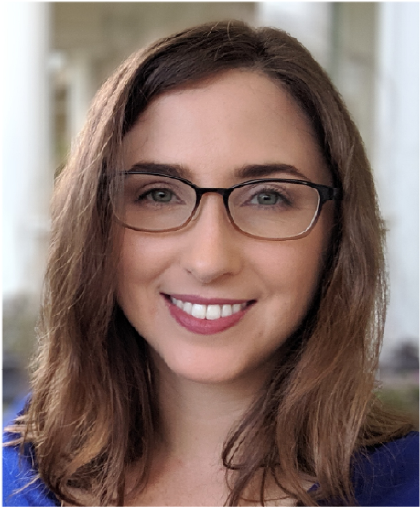 Professional headshot of a person with shoulder-length brown hair and rectangular dark-framed eyeglasses. The person is photographed from the shoulders up and is facing the camera. The individual is wearing a blue blazer over a light-colored top. The background is softly blurred, featuring neutral tones and an indoor setting with indistinct architectural elements. The image is well lit and tightly cropped around the face and upper shoulders, creating a professional portrait-style appearance.