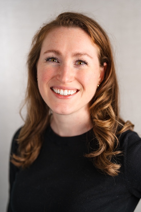 Professional head-and-shoulders portrait of a person with shoulder-length, wavy light brown hair wearing a black long-sleeved top. The person is facing the camera, and the image is cropped from the upper torso upward. The portrait is taken against a softly blurred light gray background with natural, even lighting. The photograph has a shallow depth of field that keeps the face in focus while the background remains out of focus, creating a clean and professional appearance.  Provide your feedback on BizChat