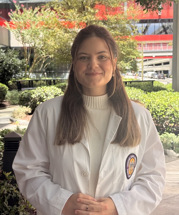 Person standing outdoors in a landscaped courtyard, wearing a white lab coat over a cream-colored ribbed turtleneck sweater. The individual has long brown hair and is photographed from about the waist up, facing the camera with hands clasped in front. A circular embroidered patch is visible on the lab coat pocket. The background includes shrubs, trees, walkways, benches, and a multi-story building with prominent red architectural features. Natural daylight illuminates the scene, with the background slightly softened while remaining recognizable.  Provide your feedback on BizChat