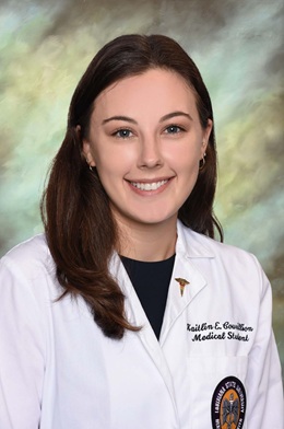 Professional studio portrait of a person wearing a white medical student lab coat over a dark shirt. The image is cropped from the chest upward, with the person facing the camera. The lab coat features embroidered text reading “Kaitlin E. Couvillon” and “Medical Student,” along with a university health sciences center patch and a caduceus lapel pin. The background is a softly blended studio backdrop in muted green, gray, and beige tones. The portrait is evenly lit and centered, creating a formal academic and professional appearance.