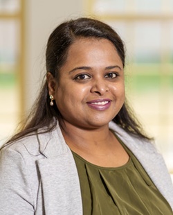 Headshot of an individual wearing a gray blazer over a green top, facing the camera against a softly blurred indoor background with warm tones.