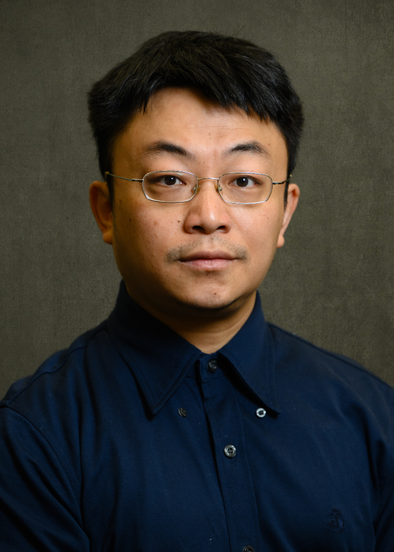 Professional head-and-shoulders portrait of a person wearing thin rectangular eyeglasses and a dark blue button-down shirt. The person is facing the camera, and the image is cropped from the upper chest upward. The background is a textured dark gray backdrop with soft, even studio lighting. The portrait is centered and formal in style, with the face in sharp focus and minimal distractions in the background.