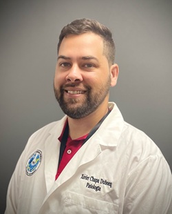 Portrait of a person wearing a white lab coat with an embroidered name and academic credentials, standing against a neutral gray background, with short dark hair and neatly trimmed facial hair, facing the camera.