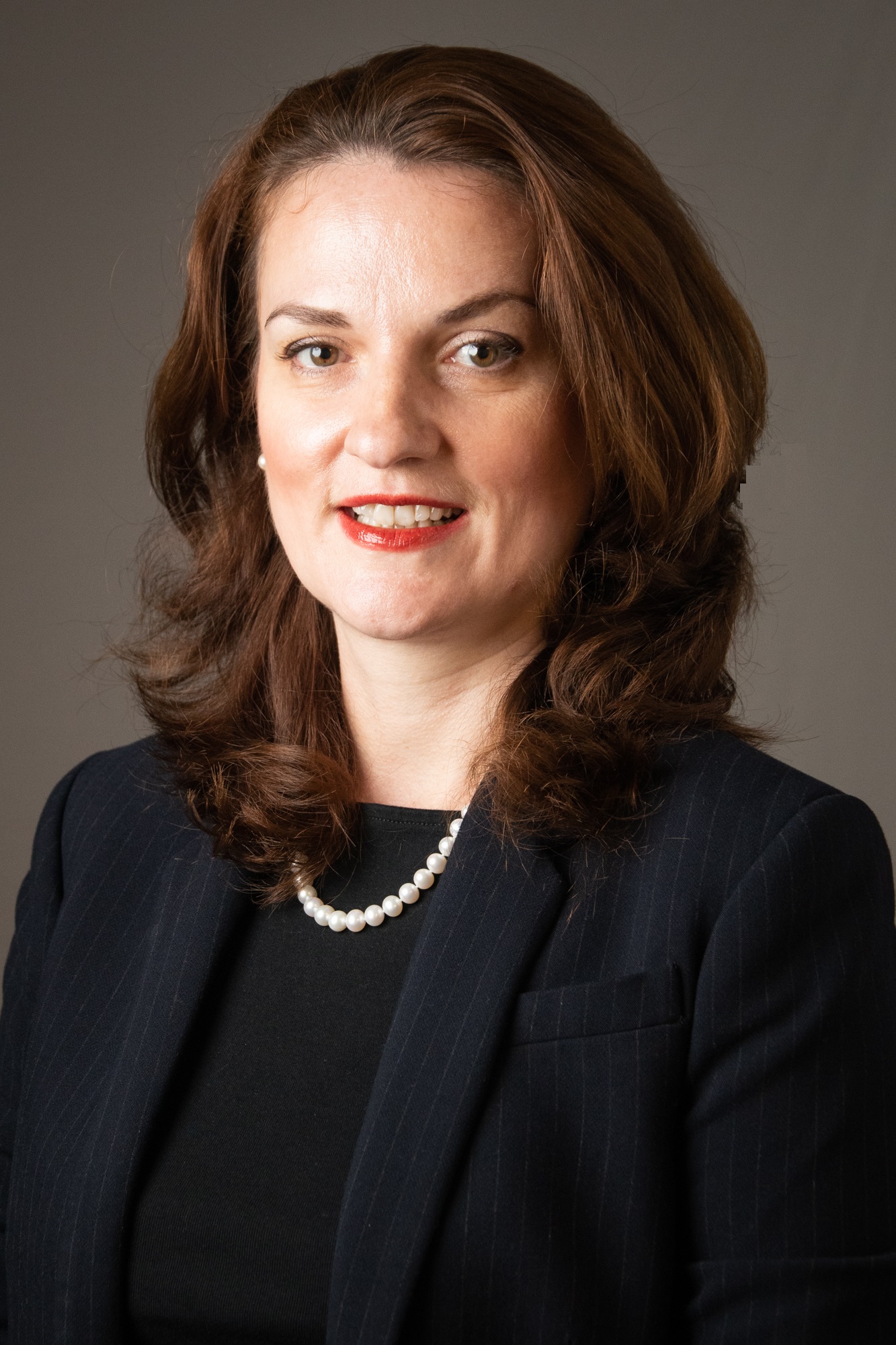 Professional head-and-shoulders portrait of a person wearing a dark pinstripe blazer over a black top and a pearl necklace. The person has medium-length brown hair styled in loose waves and is facing the camera. The background is a plain gray studio backdrop with soft, even lighting. The image is tightly cropped around the upper body and presents a formal business-style appearance.