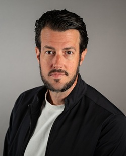Head-and-shoulders portrait of a person with short dark hair and trimmed facial hair, wearing a black jacket over a light shirt, facing the camera against a plain gray background.
