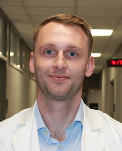 Headshot of a person with short light-brown hair wearing a white lab coat over a light blue collared shirt, facing the camera in a softly lit hallway with beige walls and ceiling lights visible in the background.