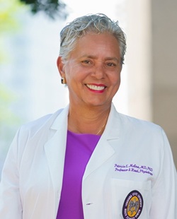 Headshot of a person with short gray hair wearing a white lab coat with an embroidered name and institutional patch over a bright purple top, facing the camera against a softly blurred outdoor or columned background.