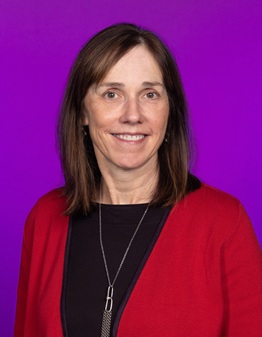 Professional head-and-shoulders portrait of a person with shoulder-length brown hair, wearing a red cardigan over a black top and a long silver necklace. The person is facing the camera against a vibrant purple gradient background, with studio lighting and no visible text or additional objects.