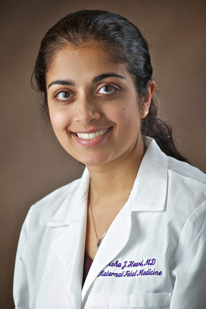Professional headshot of a person wearing a white medical coat, posed against a brown studio background.
