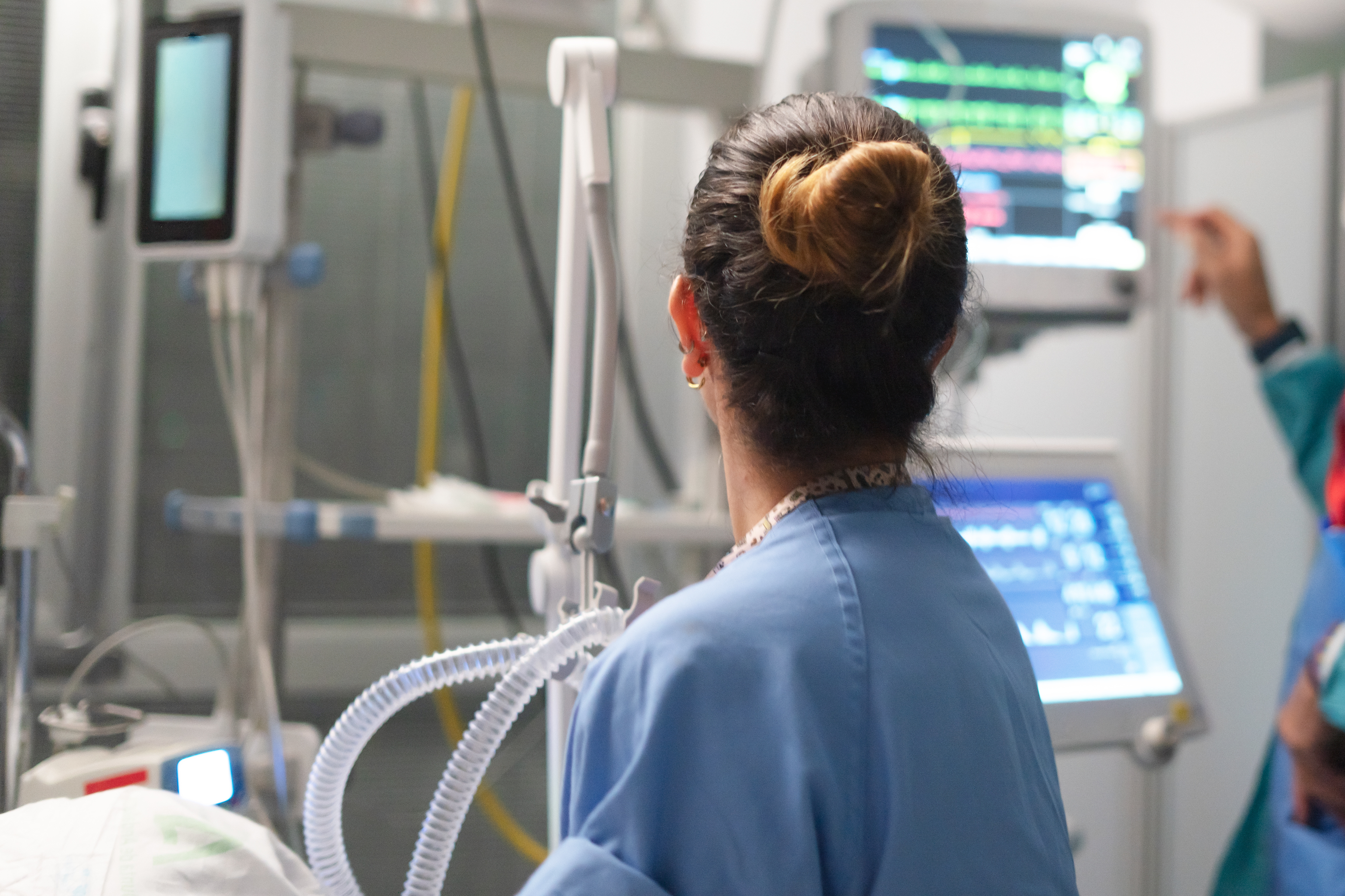 Medical professional in blue scrubs viewed from behind, looking at a patient monitor displaying vital signs in a hospital room, with ventilator tubing and medical equipment visible in the foreground.