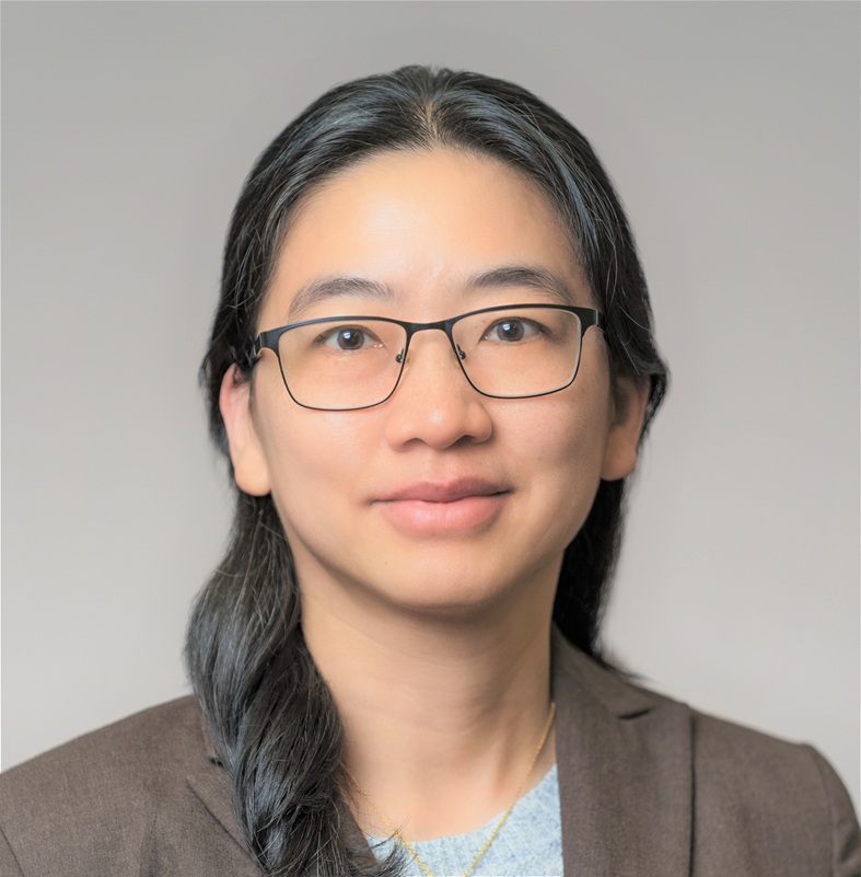 Professional head-and-shoulders portrait of a person wearing black rectangular eyeglasses, a brown blazer, and a light-colored top. The person has long dark hair worn over one shoulder and is facing the camera against a plain light gray studio background. The image is evenly lit, cropped from the shoulders up, and presented in a formal professional portrait style.  Provide your feedback on BizChat