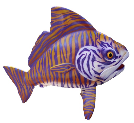 Colorful fish sculpture displayed on a concrete pedestal in an outdoor plaza surrounded by landscaping and office buildings. The sculpture is painted with bold purple and gold tiger-like stripes across its body and fins, with a large eye and detailed markings on the head. A small informational plaque sits at the base of the artwork, and trees, shrubs, and paved walkways are visible in the background.