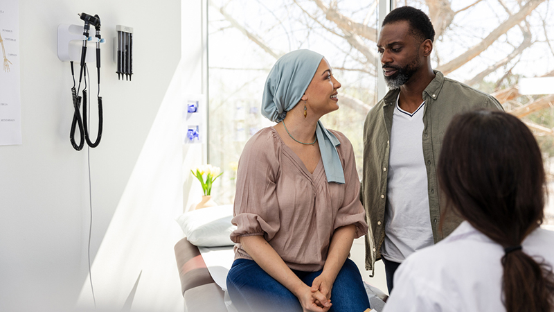 Cancer patient with headscarf smiling at supportive partner during a medical consultation.
