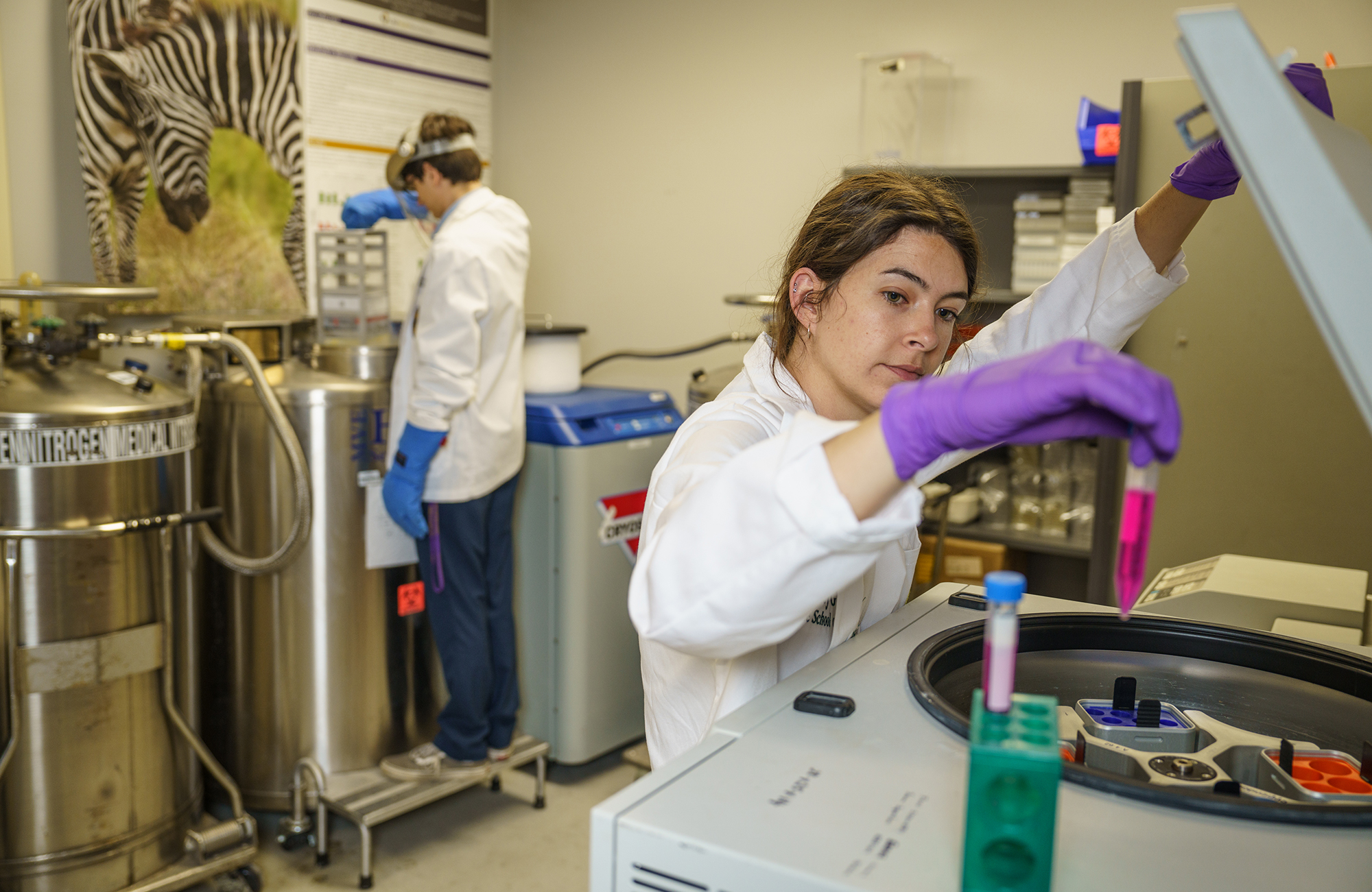 Scientists working in a laboratory equipped with cryogenic storage tanks, centrifuge, and lab equipment, wearing white lab coats, gloves, and safety gear.
