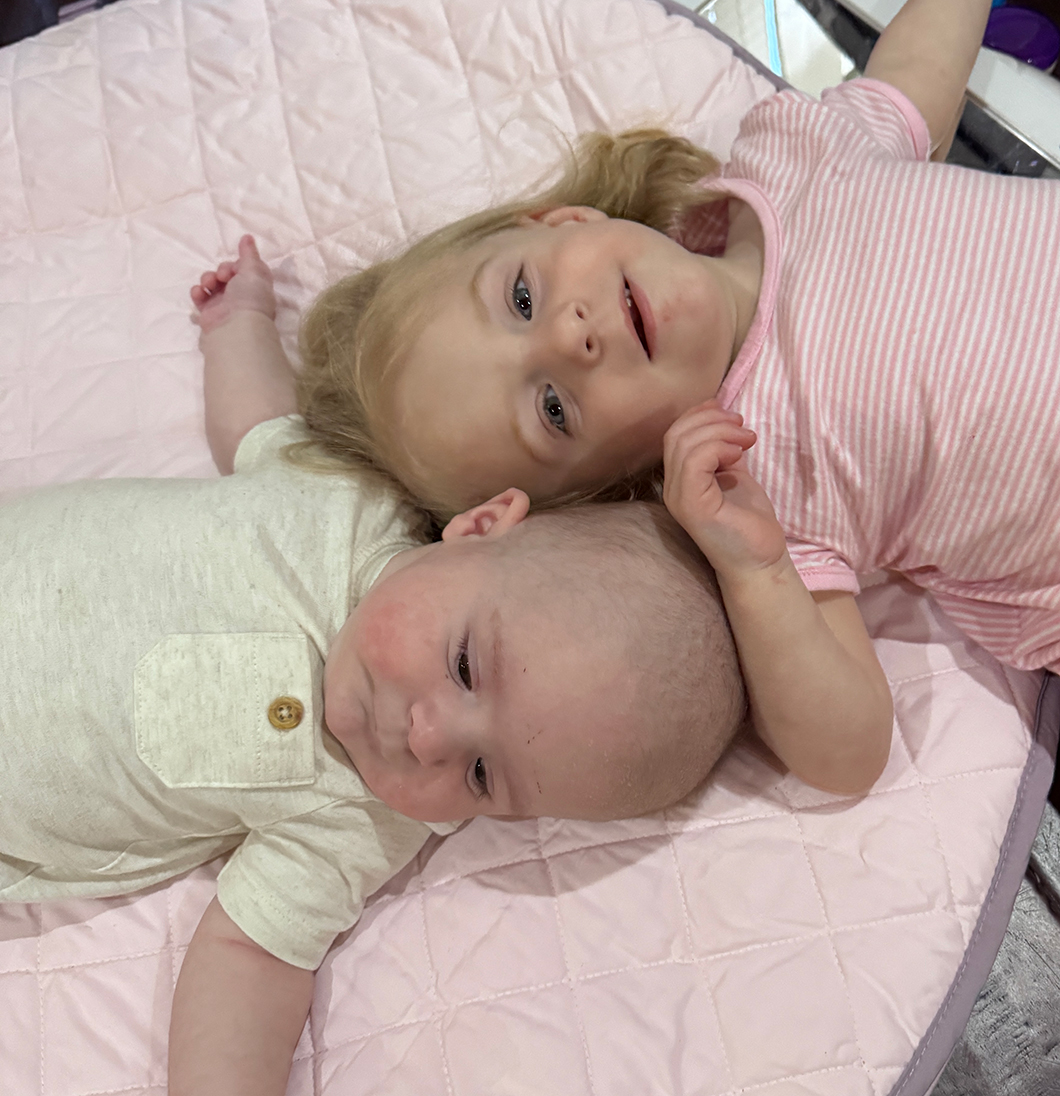 Two young siblings, Mabel and Theodore, lying on a soft pink play mat. Mabel, the older sister, smiles warmly while Theodore, the younger brother, looks curious. This image captures a moment of sibling bonding, showcasing the family’s journey after Ashley’s cancer diagnosis and fertility treatment.