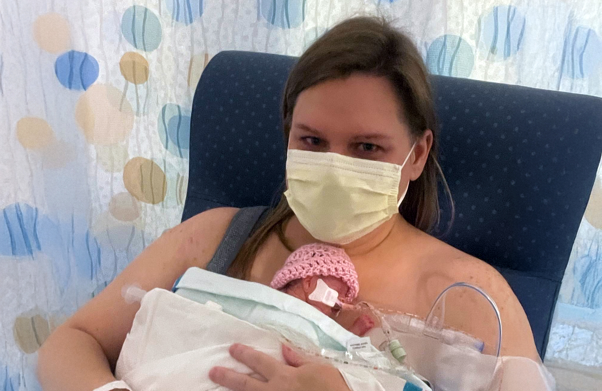 A mother, Ashley, wearing a face mask, holding her premature baby, Mabel, in a hospital setting. Mabel is wrapped in a blanket with medical equipment visible, highlighting Ashley’s journey as an endometrial cancer survivor and mother.