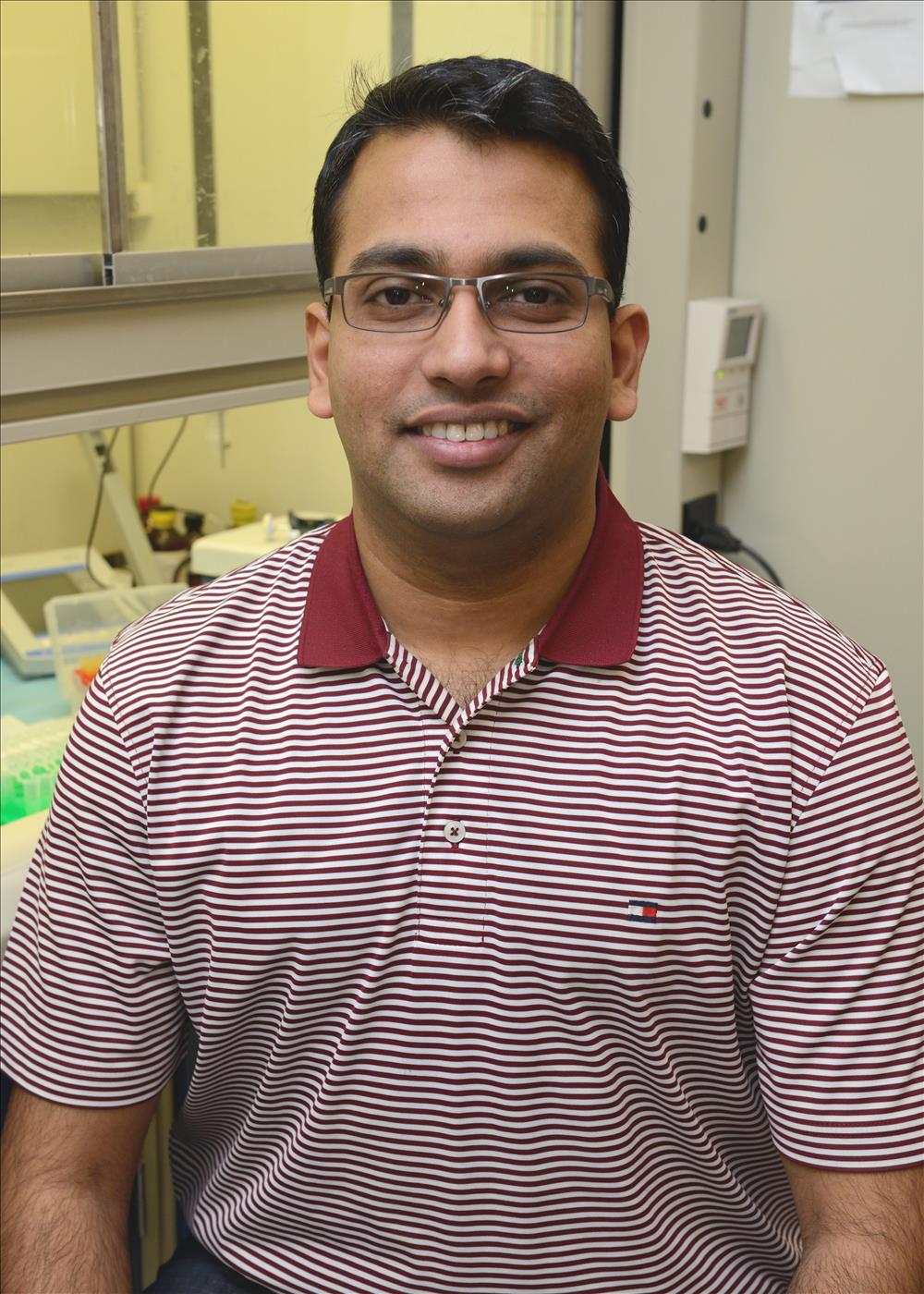 Portrait of a person wearing rectangular eyeglasses and a maroon-and-white striped collared polo shirt, seated indoors in a laboratory setting. Laboratory equipment, work surfaces, and a glass-enclosed workspace are visible in the background. The person is facing the camera, and the image is cropped from the waist up with even indoor lighting.