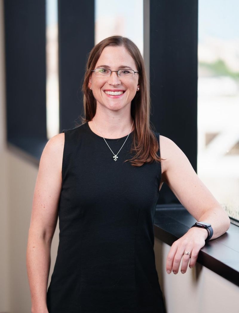 Person standing indoors beside a large window, facing the camera. The person is wearing a sleeveless black dress, a small pendant necklace, and glasses. One arm rests on the window ledge with a watch and ring visible. Vertical window frames and a blurred outdoor view appear in the background.