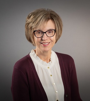 Headshot of a person wearing glasses, a white ruffled-collar blouse, and a burgundy cardigan against a gray studio background.