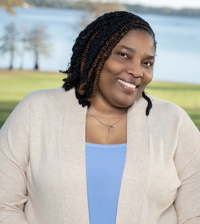 Person with braided hair wearing a light cardigan and blue top, standing outdoors near a lake with trees and grass in the background.