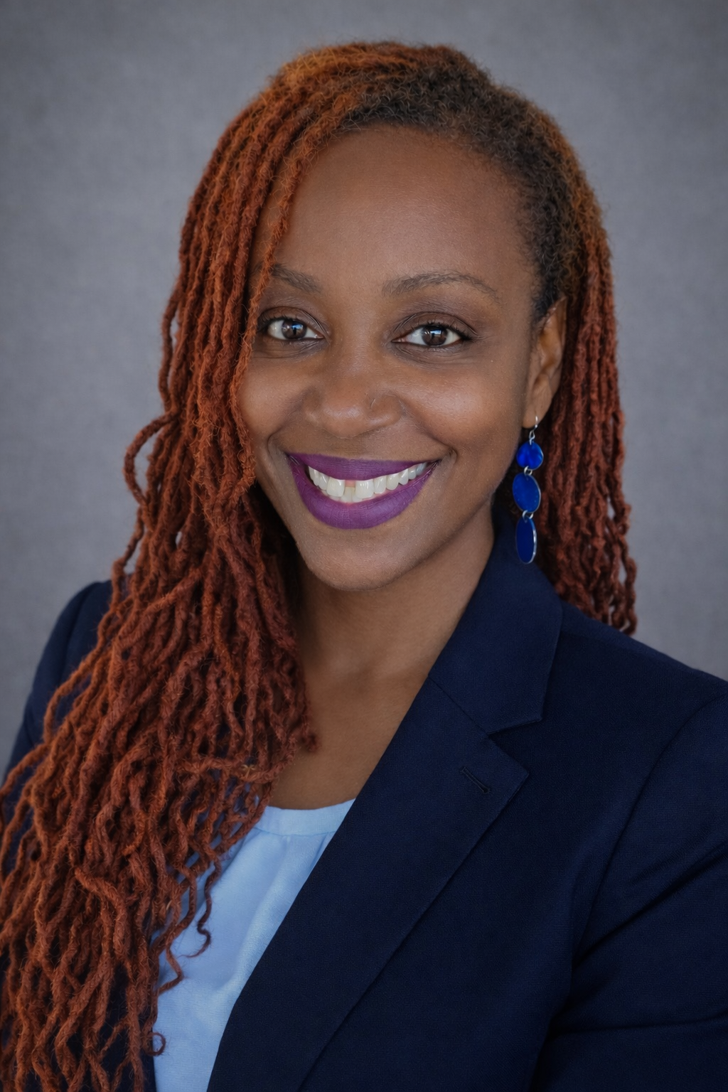 Professional headshot of a person with long reddish locs, wearing a navy blazer, white top, and blue earrings against a gray background.