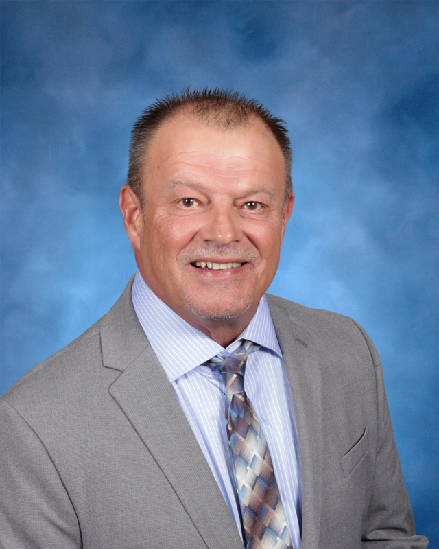 Professional headshot of a person wearing a light gray suit, striped shirt, and patterned tie against a blue studio background.