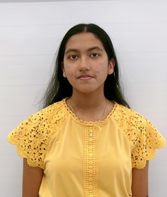 Portrait-style photo of a person standing against a plain white background, facing the camera. The person has long dark hair and is wearing a bright yellow blouse with decorative lace cap sleeves and a lace-trimmed front panel. The image is cropped from the upper torso upward, with even lighting and no visible accessories or background objects.