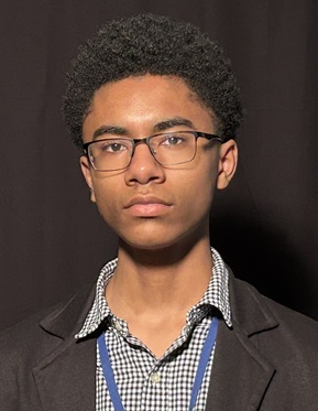 Head-and-shoulders portrait of a person with short, dark, hair wearing rectangular eyeglasses, a dark blazer, and a patterned collared shirt. The person is facing the camera against a dark studio background with focused lighting on the face.