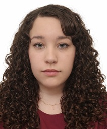 Head-and-shoulders portrait of a person with long, dark, tightly curled hair wearing a burgundy top. The person is facing the camera against a plain light gray background with even studio lighting.