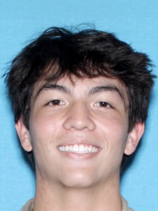 Close-up portrait of a person with short, dark, tousled hair facing the camera against a solid blue background. The image is tightly cropped around the head and upper shoulders. The person is wearing a light-colored collared shirt and is photographed with even lighting, similar to an identification or school portrait style.
