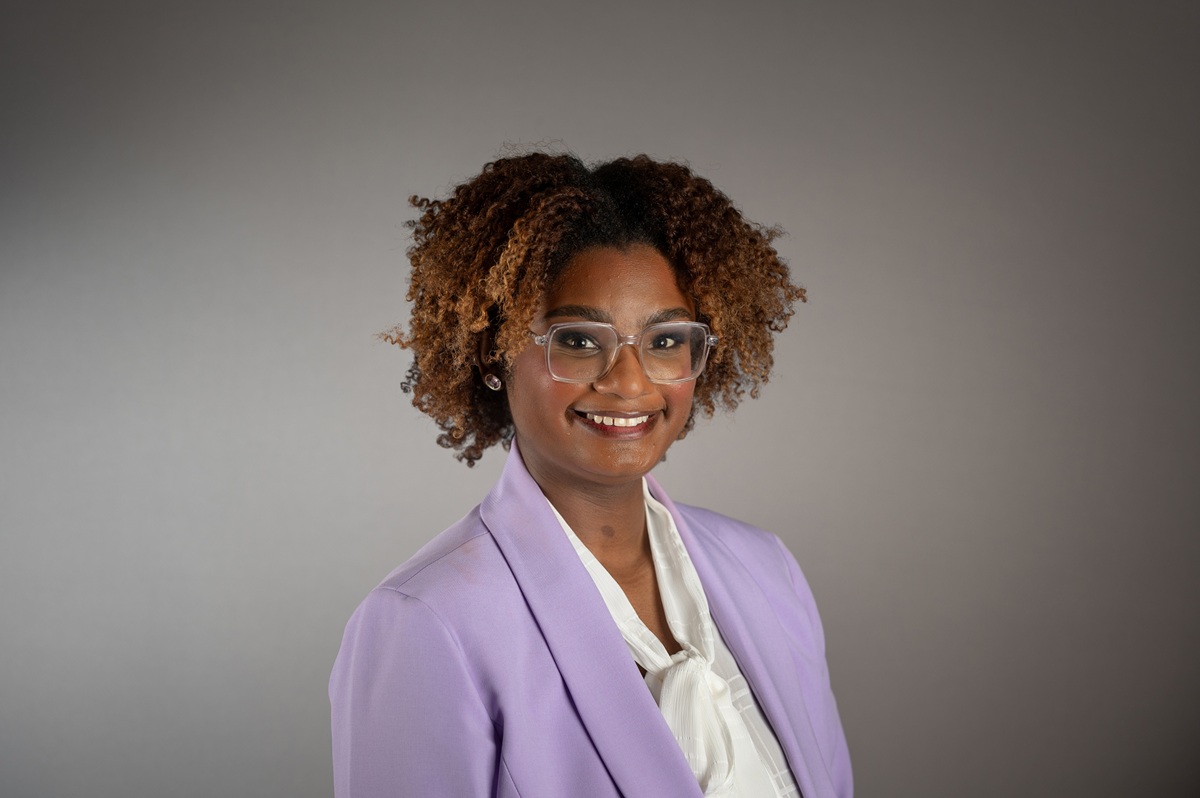 A person with curly brown hair, clear-framed glasses, and a light purple blazer poses against a plain gray backdrop. They wear a white blouse with a bow at the collar and look directly at the camera with a neutral, professional expression.