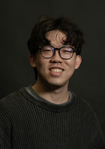 Professional studio portrait of a person with short, dark wavy hair wearing round eyeglasses and a dark textured crew-neck sweater. The person is facing the camera against a dark gradient studio background. The image is framed from the chest up and lit with soft, focused studio lighting.