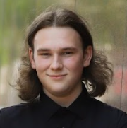Head-and-shoulders outdoor portrait of a person with shoulder-length light brown hair wearing a black collared shirt. The person is facing the camera with a blurred background of greenery and natural colors. The image is evenly lit with natural daylight and framed closely around the face and shoulders.