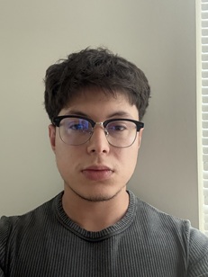 Head-and-shoulders portrait of a person with short dark hair wearing rectangular eyeglasses and a dark ribbed crew-neck shirt. The person is facing the camera indoors, positioned near a window with vertical blinds visible along the right side of the image. The background is a plain light-colored wall, and the image is lit by soft natural light from the window.
