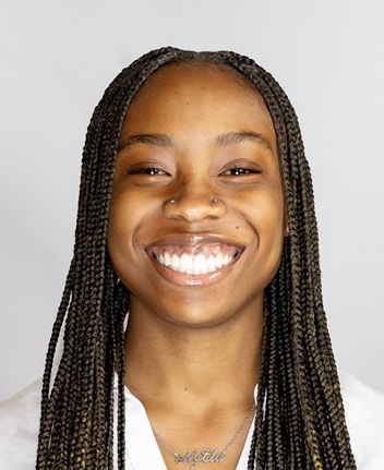 Professional head-and-shoulders portrait of a person with long box braids wearing a light-colored top and a nameplate necklace. The person is facing the camera against a plain light gray studio background. The image is closely cropped around the face and shoulders and lit with bright, even studio lighting.